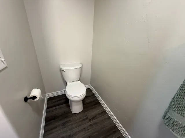 White toilet with chrome flush lever, dark wood-finish flooring, and white baseboards