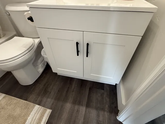 White shaker-style vanity with matte black hardware, white countertop, and dark wood-finish flooring