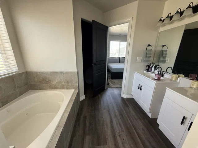 Soaking tub with tile surround, wood-finish flooring, dual vanity with white cabinetry, and a large wall mirror