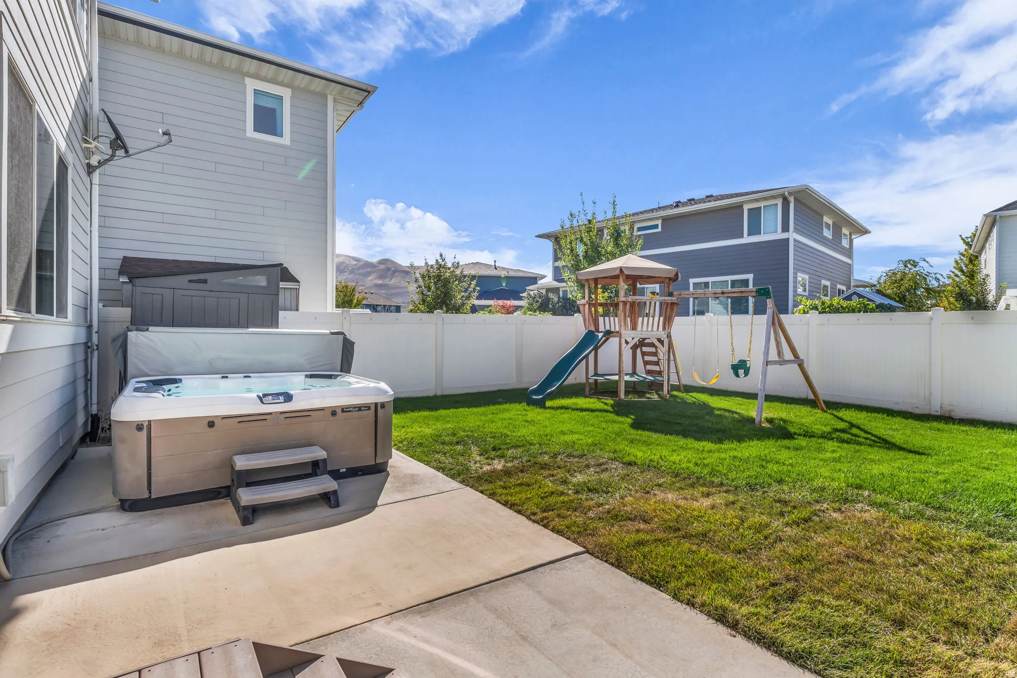 Concrete patio featuring a hot tub with steps, expansive green lawn, and white vinyl fencing
