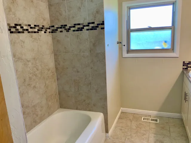 Bathtub with neutral-toned wall tiles, decorative mosaic accent band, and a window with white trim