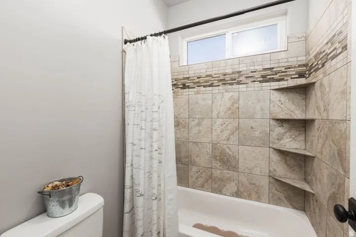 Tub and shower combination featuring tiled surround, mosaic tile accent, built-in corner shelving, and an upper window