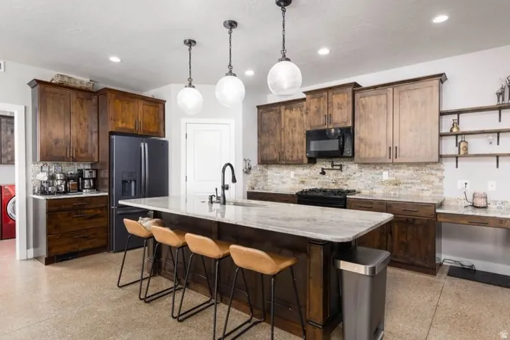 Kitchen featuring a large central island with seating, dark wood cabinetry, light stone countertops, a stone tile backsplash, and dark-finish appliances