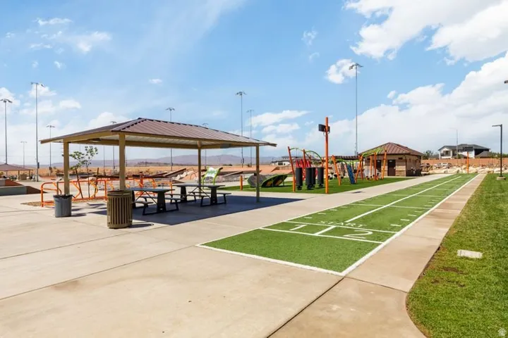 Shaded seating area with picnic tables, artificial turf shuffleboard court, built-in playground structure, and an adjacent building