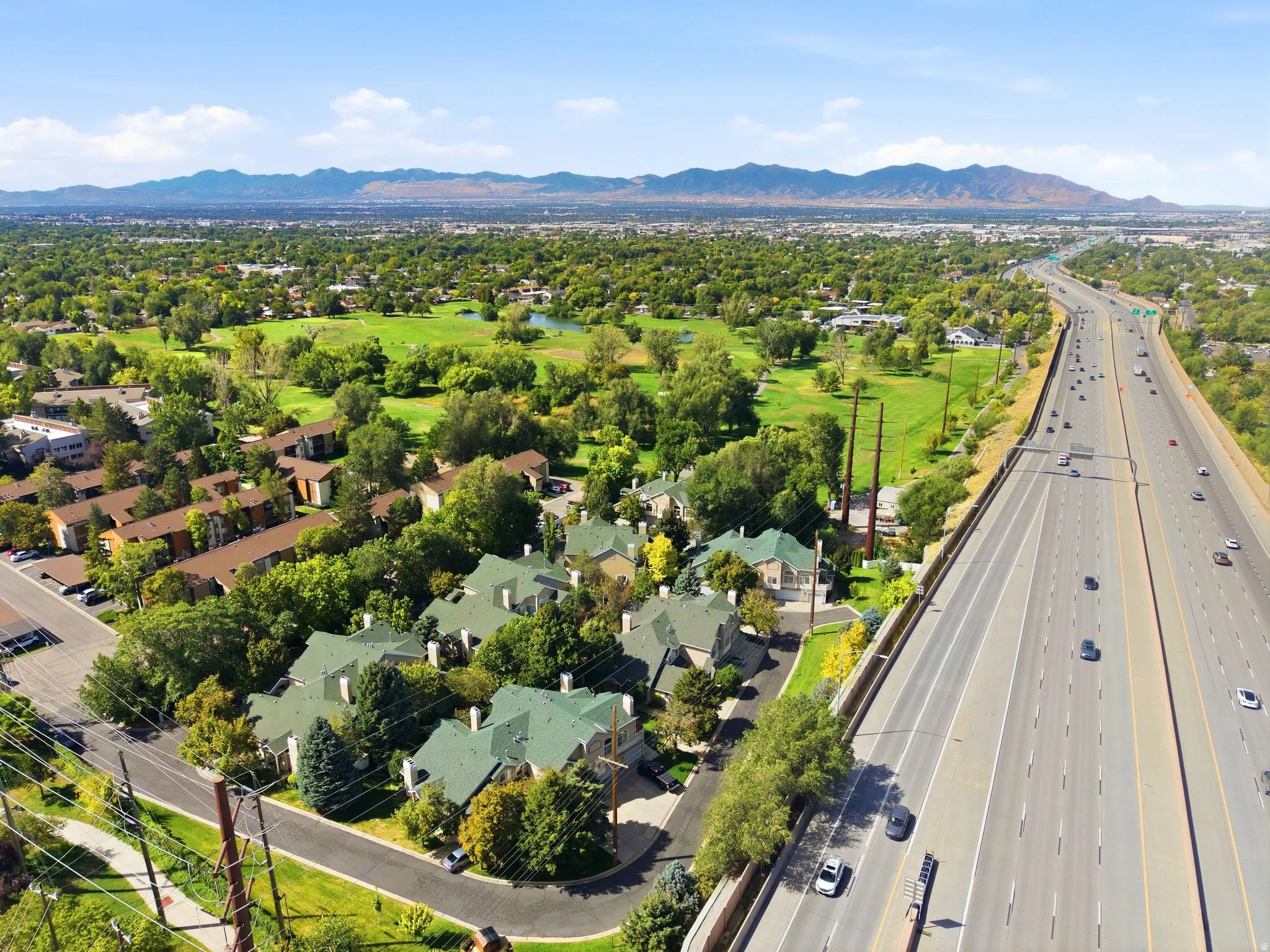 Aerial perspective showcasing a cluster of townhomes with pitched green roofs, abundant mature trees, a multi-lane highway, a sprawling green golf course, and distant mountains