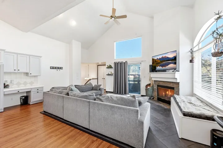 Vaulted ceilings with a ceiling fan, a fireplace with a stone surround, wood-finish flooring, built-in cabinetry with a desk, and an arched window