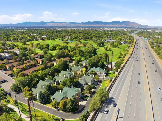 Aerial perspective showcasing a cluster of townhomes with pitched green roofs, abundant mature trees, a multi-lane highway, a sprawling green golf course, and distant mountains