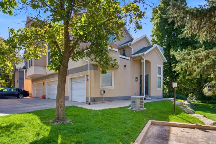 Stucco exterior with multiple garage doors, manicured lawn, mature trees, and an exterior light fixture