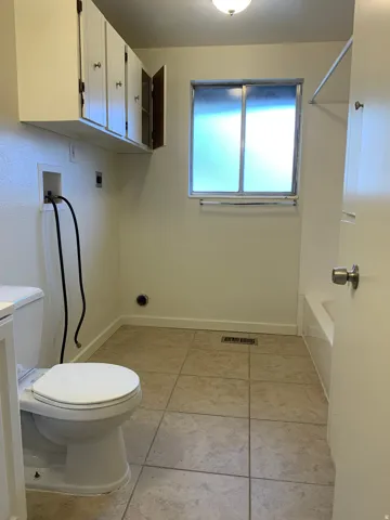 Tile flooring, white wall-mounted cabinetry, frosted window, built-in bathtub, and overhead light fixture