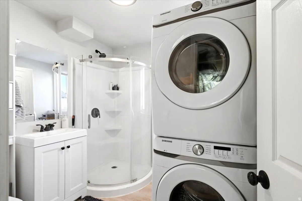 White stacked washer and dryer unit, corner shower with clear glass enclosure, white vanity with integrated sink, black faucet, and frameless mirror