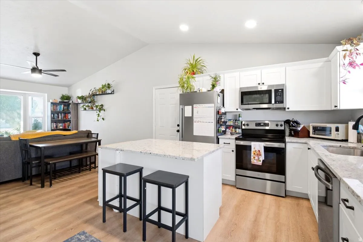 Vaulted ceiling, wood-finish flooring, white cabinetry with dark hardware, stainless steel appliances, and a kitchen island with a light-toned countertop