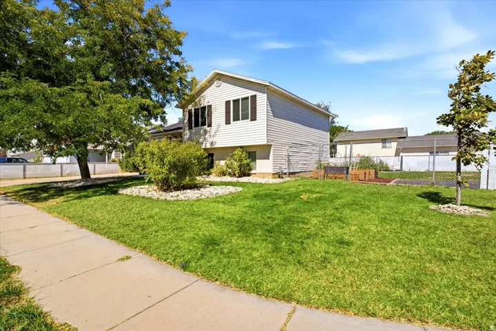 Light-colored siding exterior, dark window shutters, established front lawn, mature trees, and a concrete sidewalk