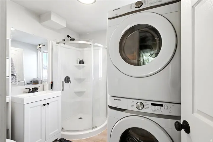 White stacked washer and dryer unit, corner shower with clear glass enclosure, white vanity with integrated sink, black faucet, and frameless mirror