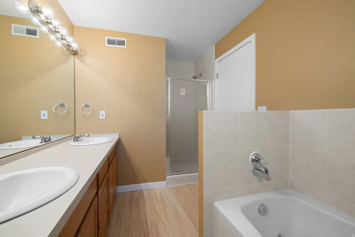 Dual vanity with integrated sinks, ample counter space, wood-finish flooring, tiled soaking tub, and a separate glass-enclosed shower