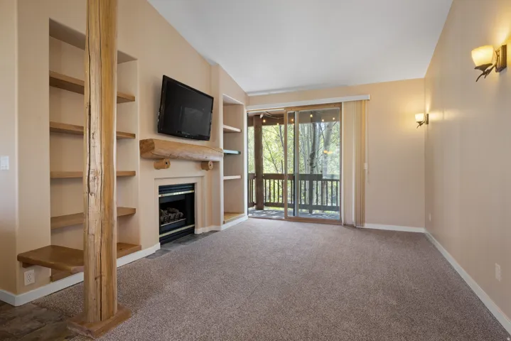 Vaulted ceiling, built-in shelving, stone tile fireplace surround with wooden mantel, sliding glass door, and outdoor balcony with railing