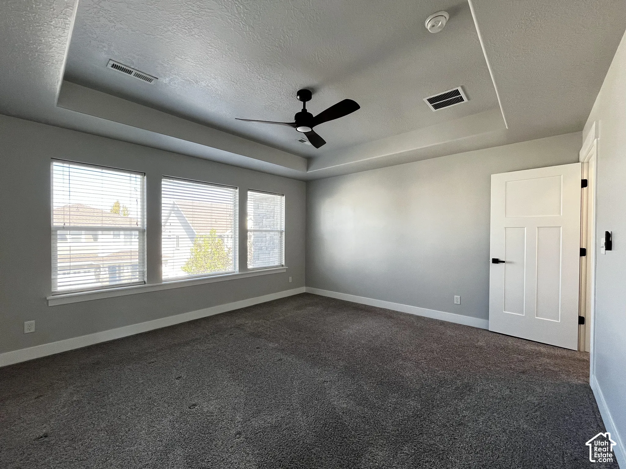 Tray ceiling with ceiling fan, three windows with blinds, gray wall paint, and gray carpet flooring