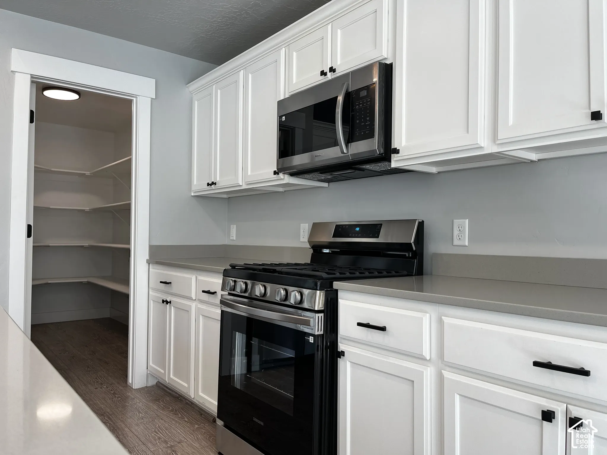 White shaker cabinetry with matte black hardware, stainless steel microwave, stainless steel range, gray countertops, and a walk-in pantry with wood-finish flooring