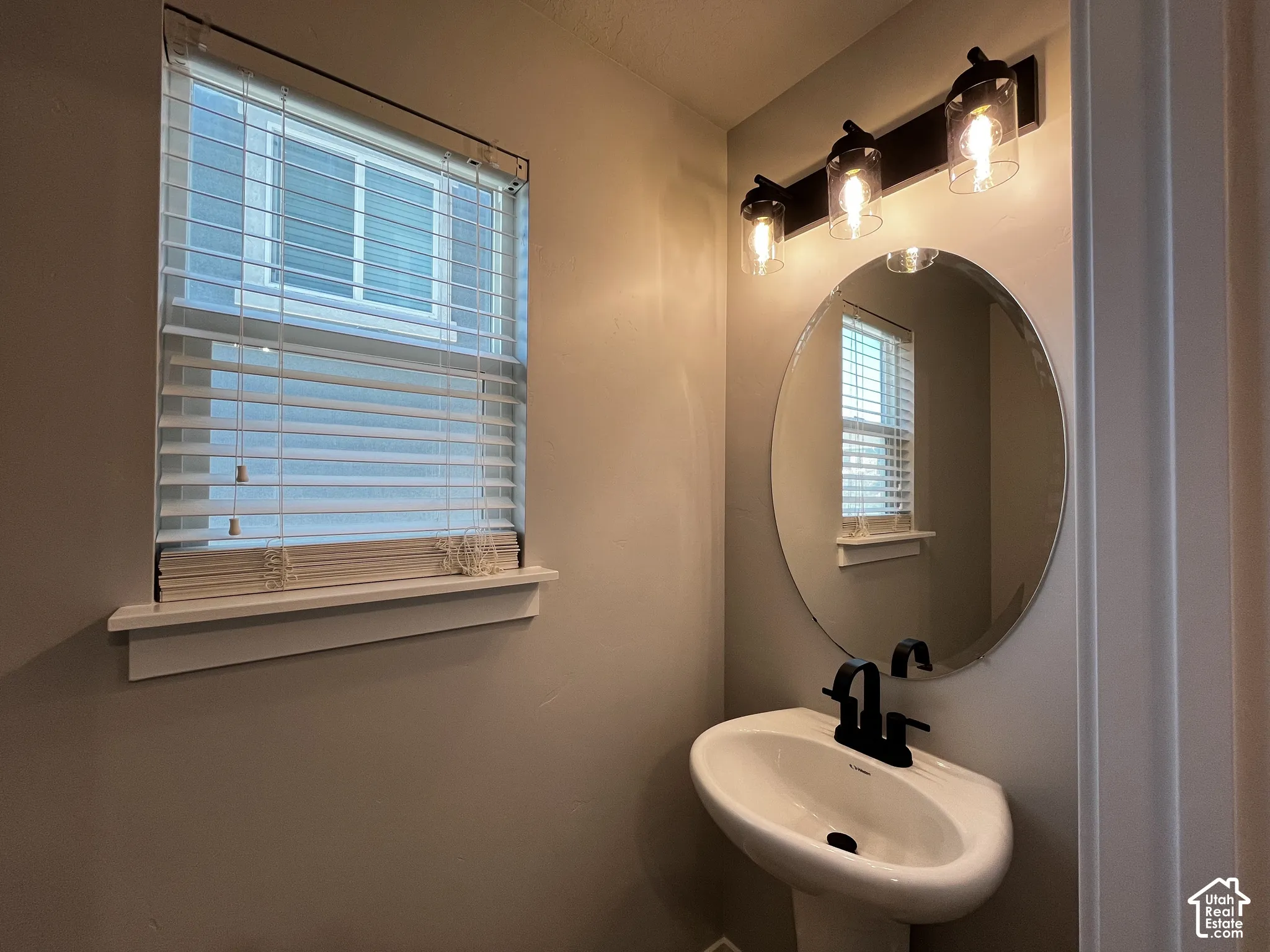 Pedestal sink with matte black faucet, oval mirror, and three-light vanity fixture