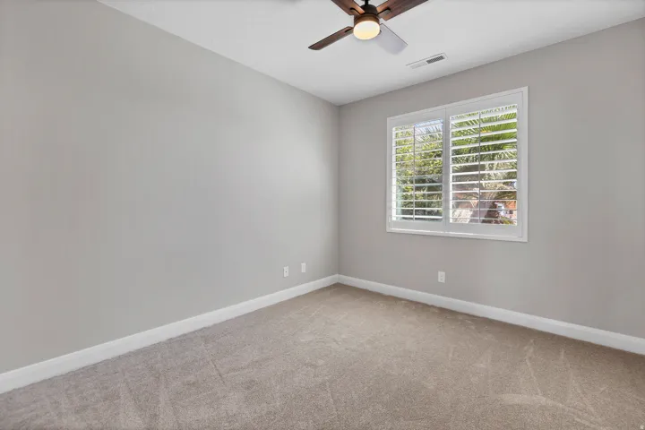 Neutral wall color, plush carpet, window with white plantation shutters, ceiling fan with light fixture
