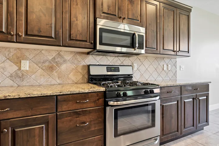 Dark wood-finish cabinetry with crown molding, stainless steel microwave, stainless steel range, diamond-patterned tile backsplash, and stone countertops