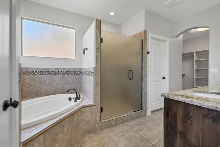 Soaking tub with tile surround, walk-in shower with frosted glass enclosure, single vanity with stone countertop, and built-in closet shelving
