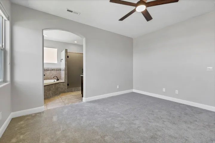 Light gray wall paint, gray carpet flooring, ceiling fan with light fixture, arched doorway, and built-in tub with tile surround