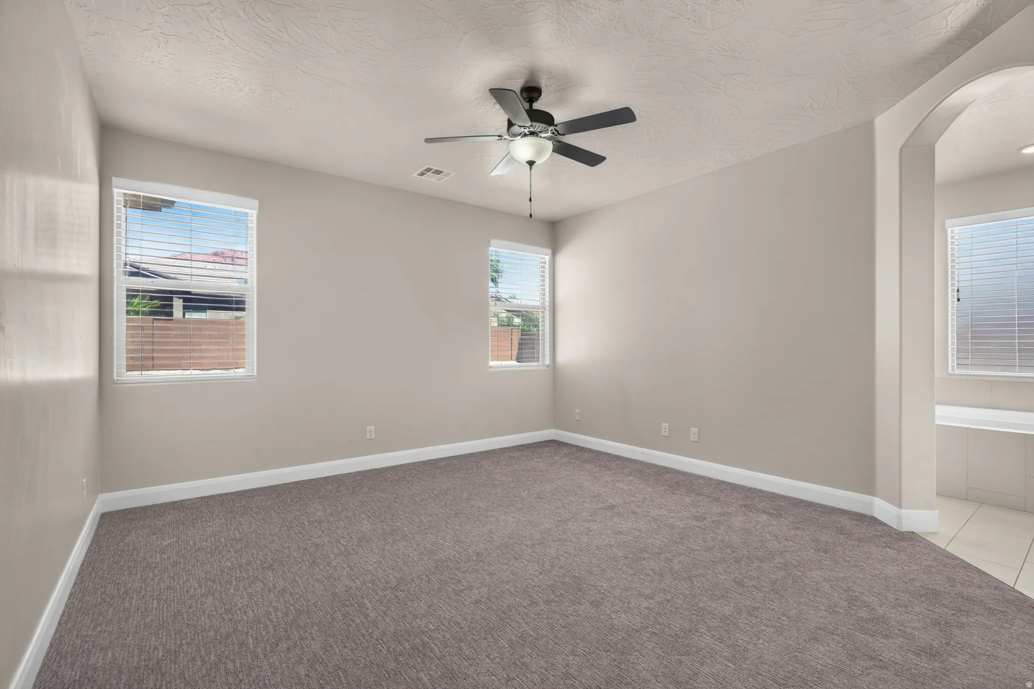 Neutral tone wall color, textured ceiling, ceiling fan with integrated lighting, carpeted flooring, and an arched entry