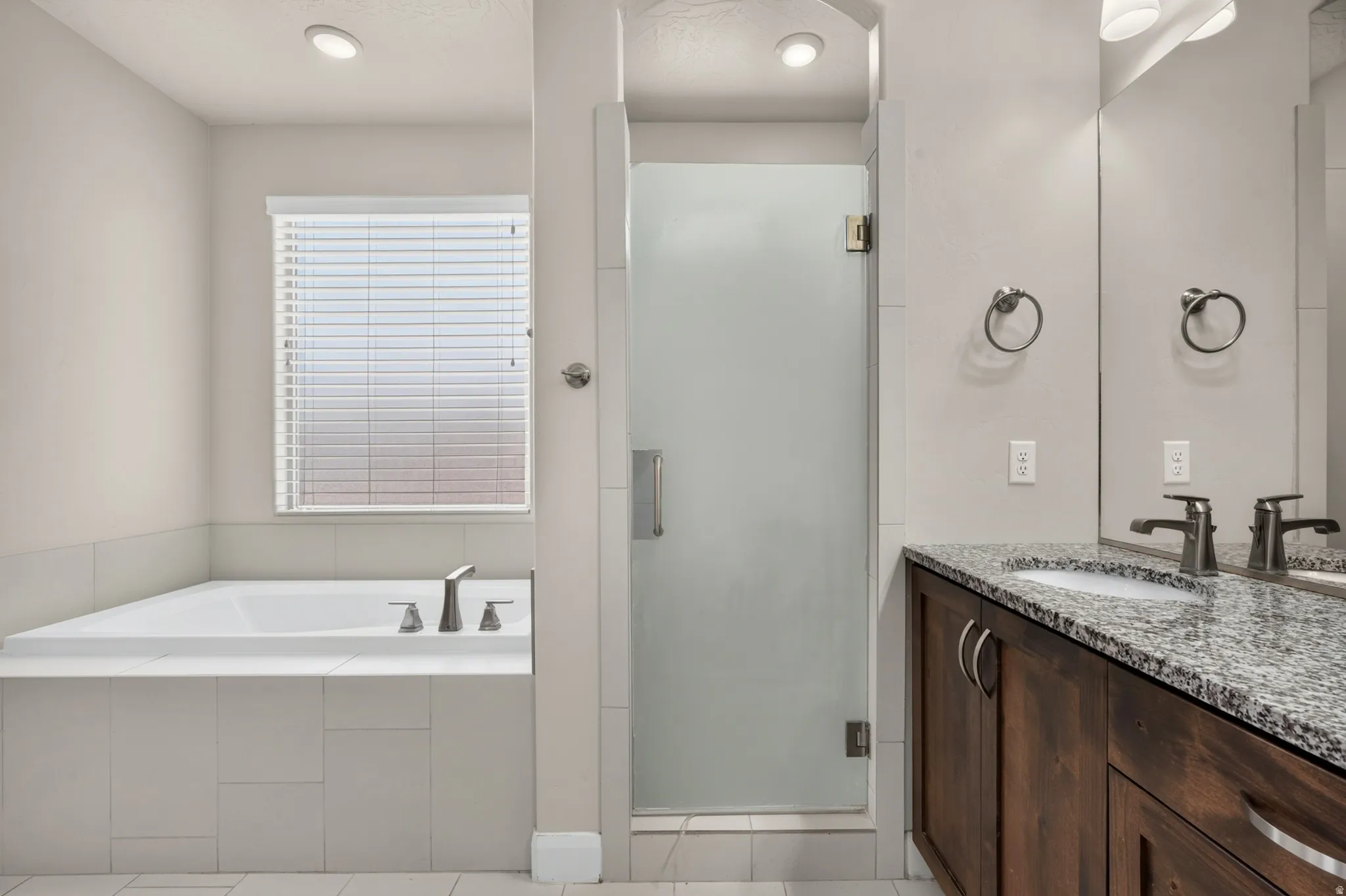 Built-in bathtub, glass-enclosed shower, dual vanity with granite countertop, wood-finish cabinetry, and recessed lighting