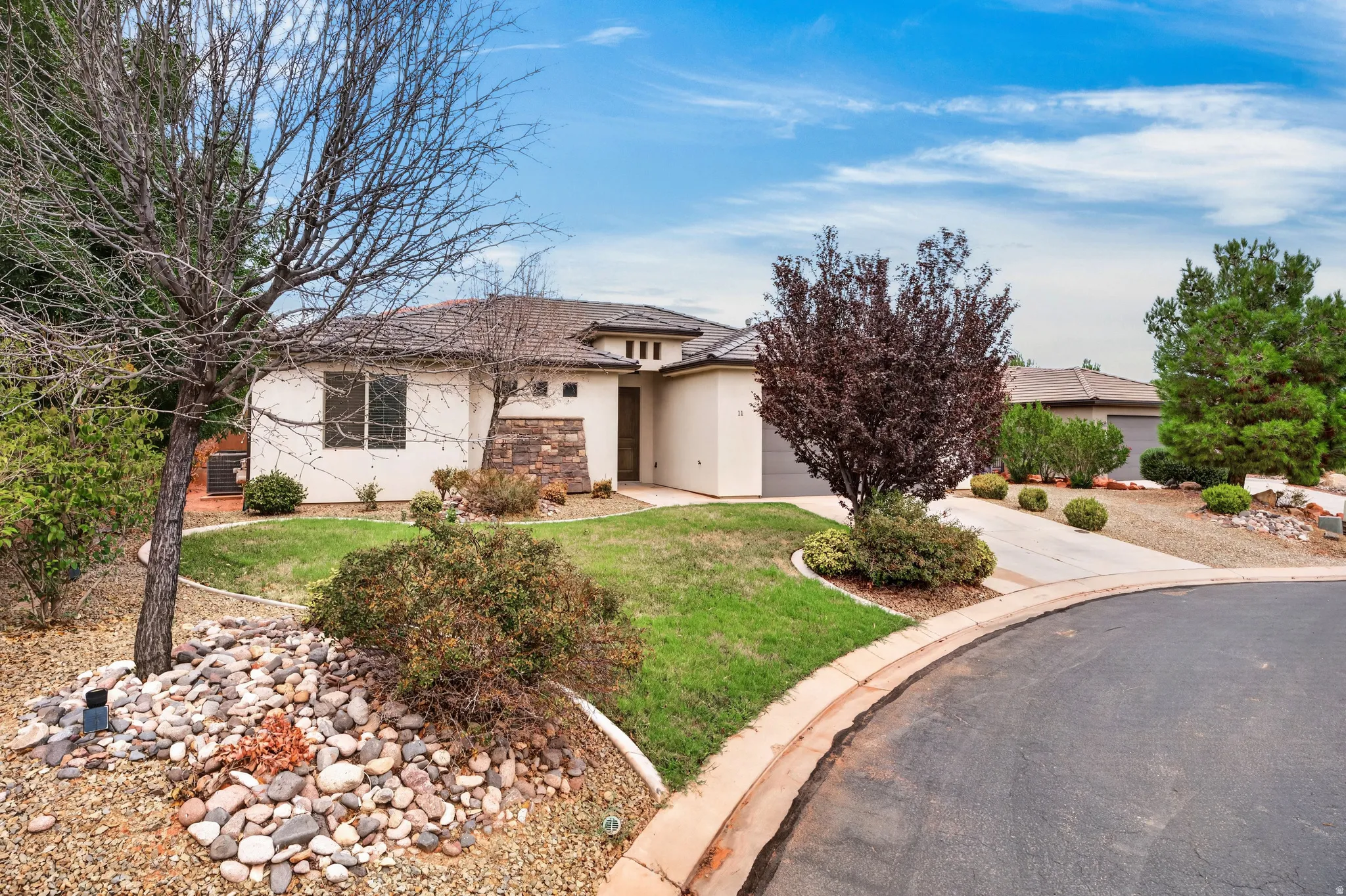 Stucco exterior with stone accents, a landscaped front yard with mature plantings, and a paved driveway