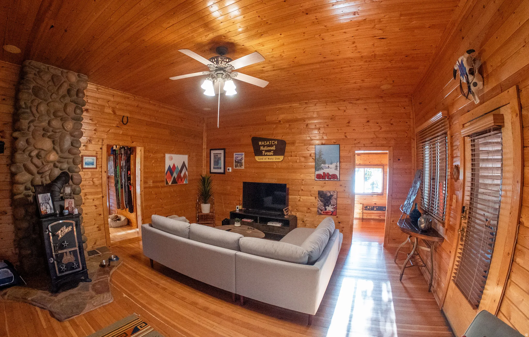 Wood-paneled walls and ceiling, stone-clad fireplace, wood-finish flooring, and a ceiling fan with integrated lighting