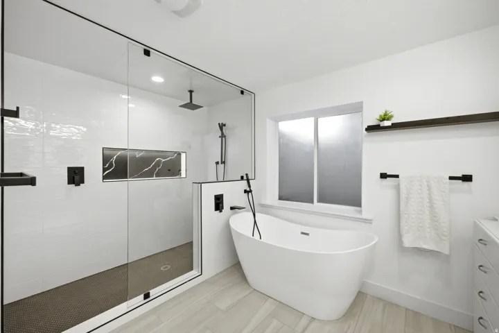Freestanding soaking tub, frameless glass shower enclosure with matte black fixtures, integrated shower niche with black veined surface, white subway tile shower surround, and wood-finish flooring