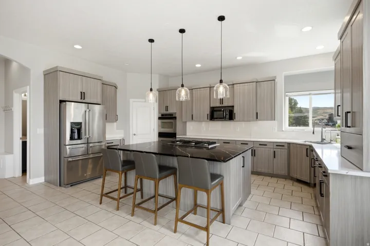 Kitchen featuring stainless steel appliances, light wood-finish cabinetry with black hardware, a center island with black countertop, white tile flooring, and pendant lighting