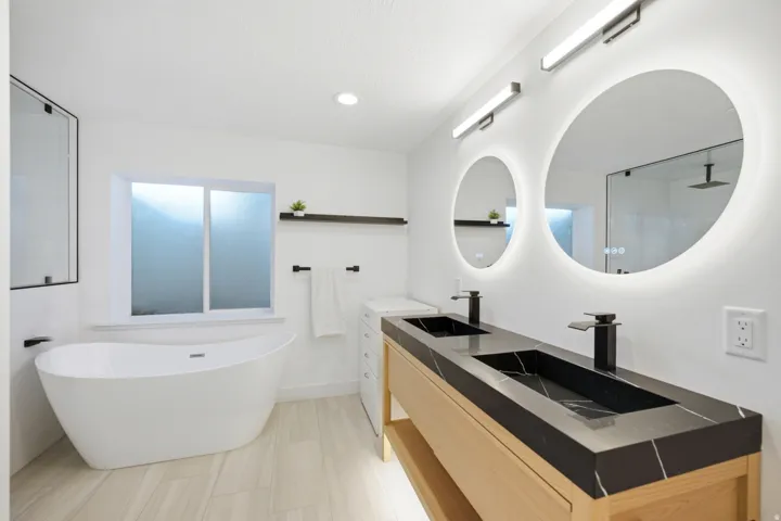 Freestanding soaking tub, dual illuminated vanity mirrors, matte black fixtures, wood-finish flooring, and a stone-finish dual sink countertop