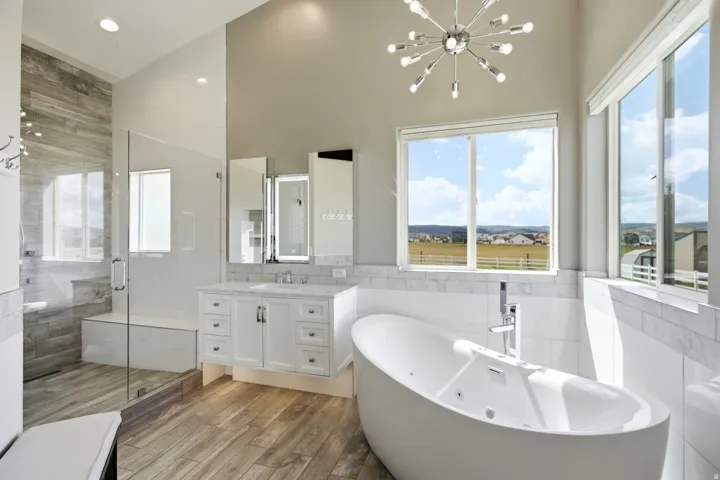 Freestanding soaking tub, glass-enclosed shower with bench seating, floating vanity with dual illuminated medicine cabinets, wood-finish flooring, and a contemporary light fixture