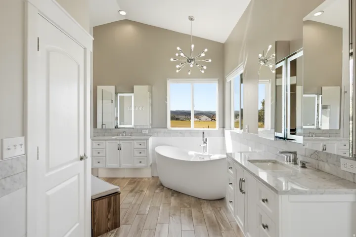 Vaulted ceiling with a contemporary chandelier, freestanding soaking tub, dual vanities with undermount sinks, wood-finish flooring, and a large window