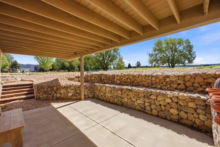 Covered concrete patio with exposed wood ceiling beams, stone gabion retaining walls, and gravel landscaping
