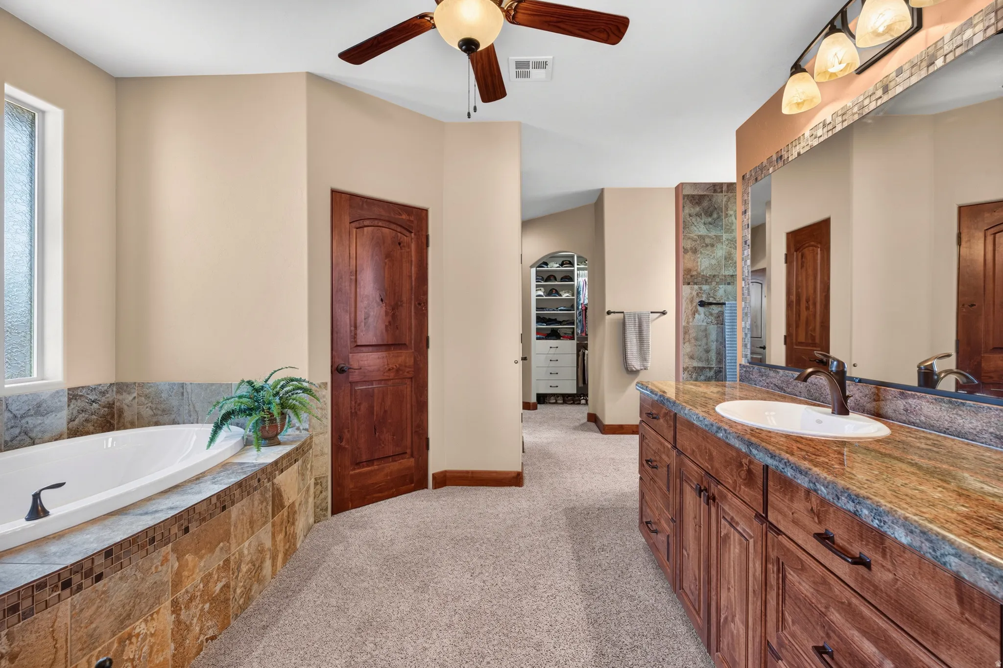 Soaking tub with tiled surround, wood-finish vanity with countertop, ceiling fan, and built-in closet storage