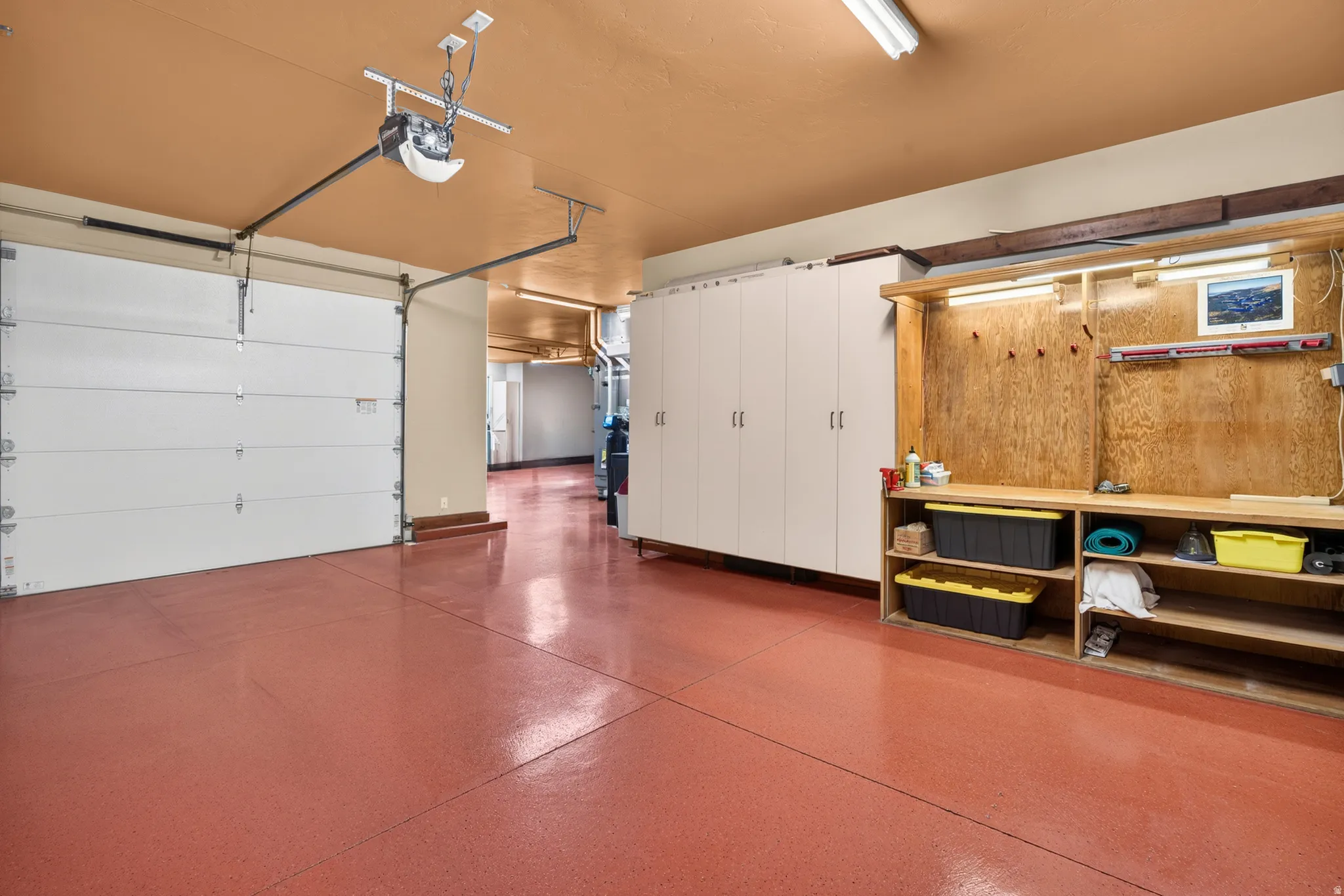 Spacious garage featuring a durable red-finish floor, overhead fluorescent lighting, a built-in workbench with shelving, and extensive white-paneled storage cabinetry