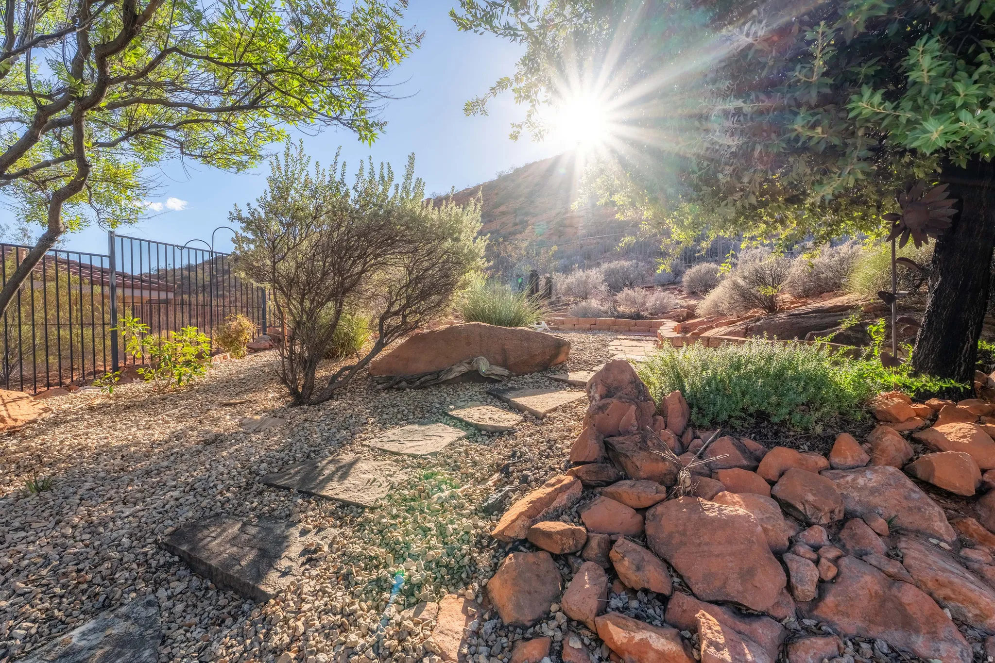 Landscaped exterior featuring a metal fence, natural stone hardscaping, mature trees, and drought-tolerant plantings
