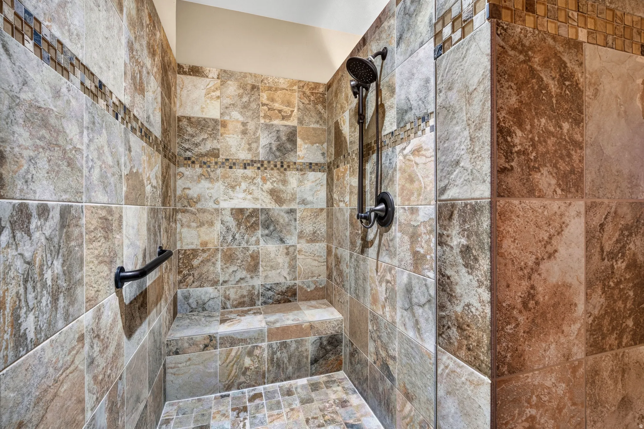 Walk-in shower with natural stone-look tile, mosaic tile accent, built-in bench, and black grab bar