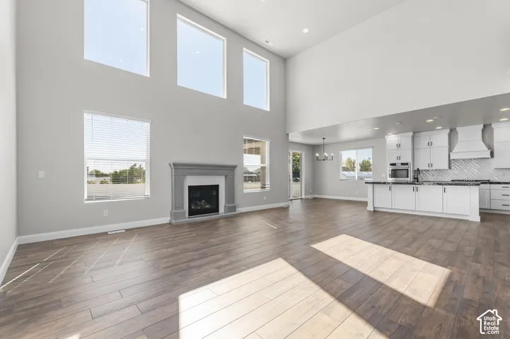 High-ceilinged living area featuring wood-finish flooring, a fireplace with a decorative mantle, oversized windows, and a kitchen equipped with white cabinetry and an island