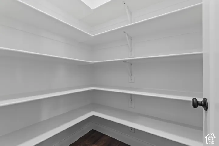 Walk-in storage space featuring wrap-around white shelving, wall-mounted support brackets, and dark wood-finish flooring