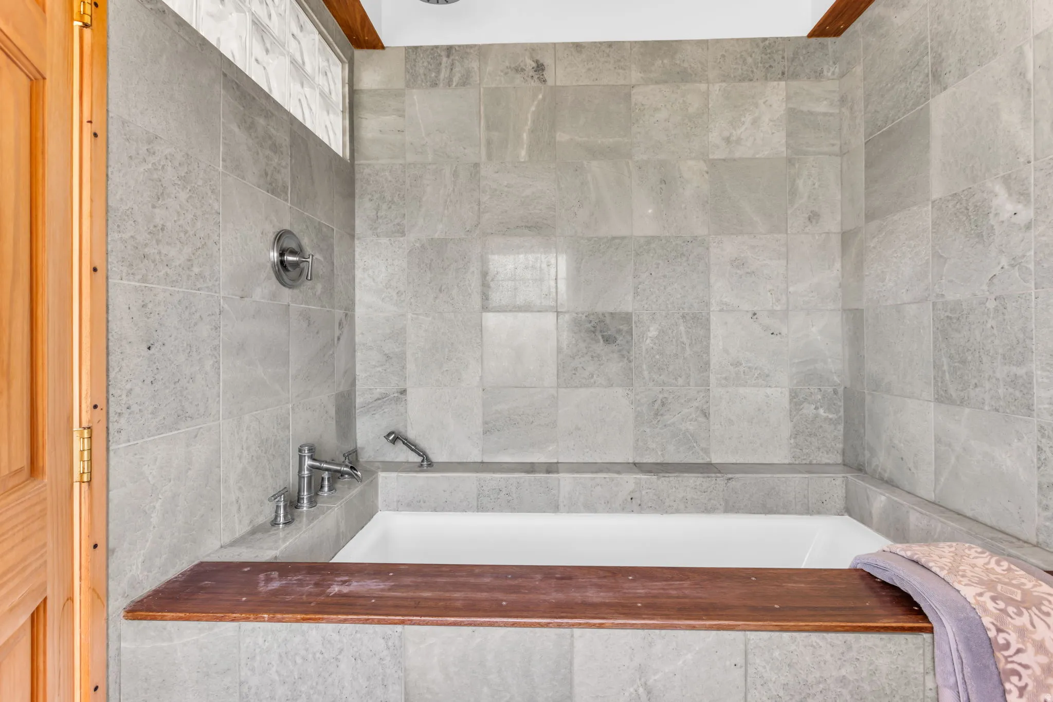 Primary bathroom with built-in soaking tub with a wood-finish deck, surrounded by large format gray wall tile, and polished chrome fixtures