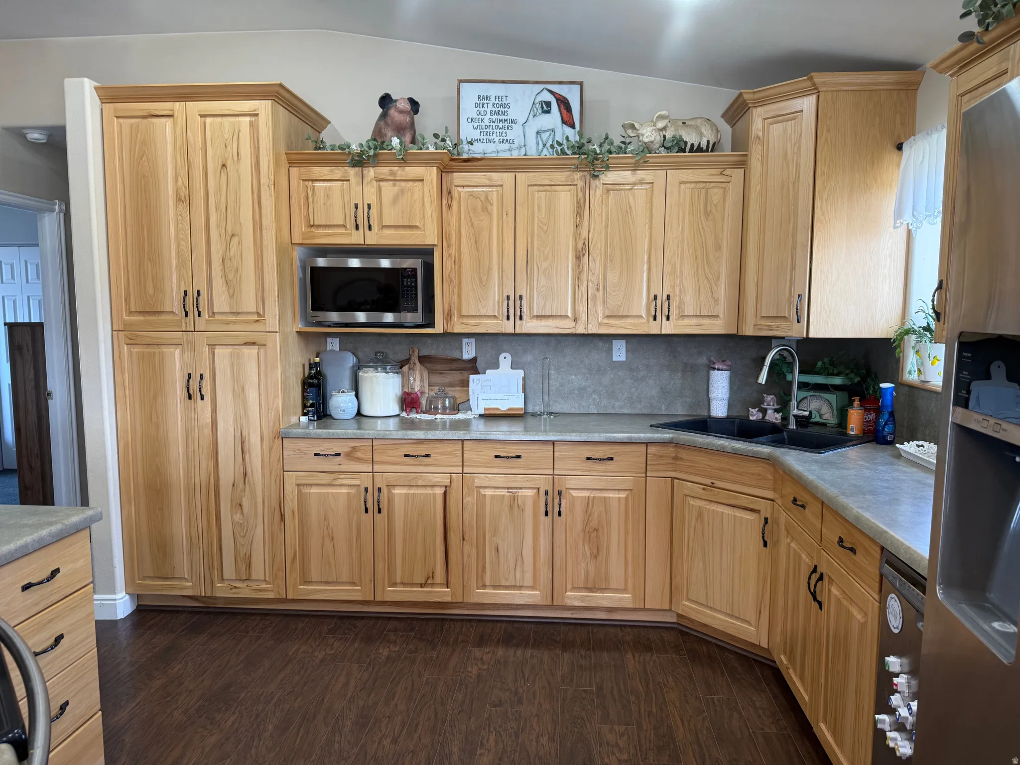 Wood-finish flooring, light wood cabinetry, stainless steel microwave, black sink fixture, and a tile backsplash