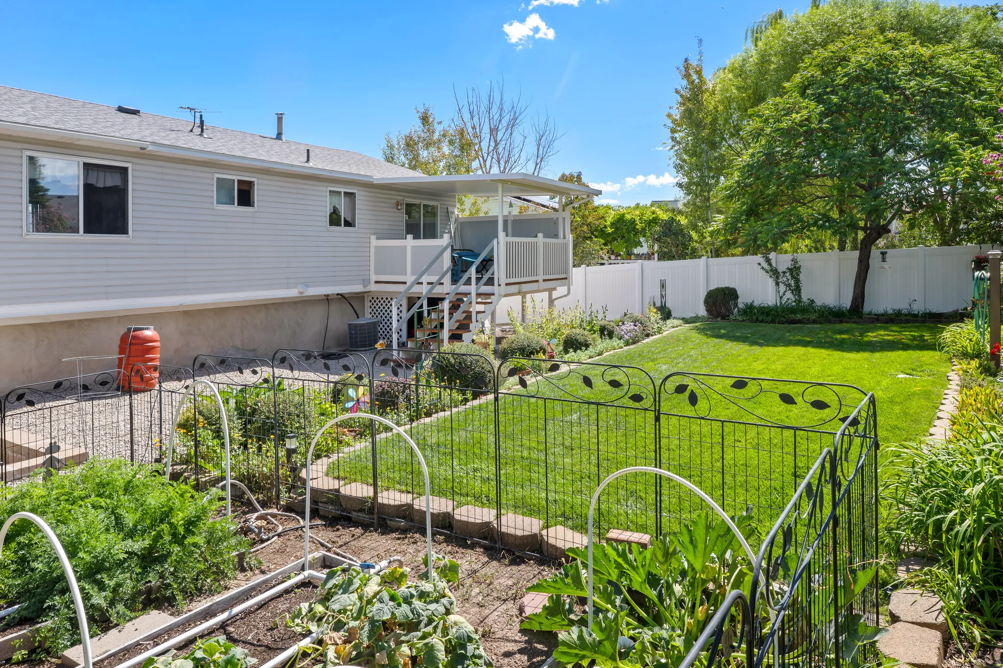 Expansive backyard featuring a manicured lawn, established garden beds, white privacy fencing, and a built-in deck with stairs