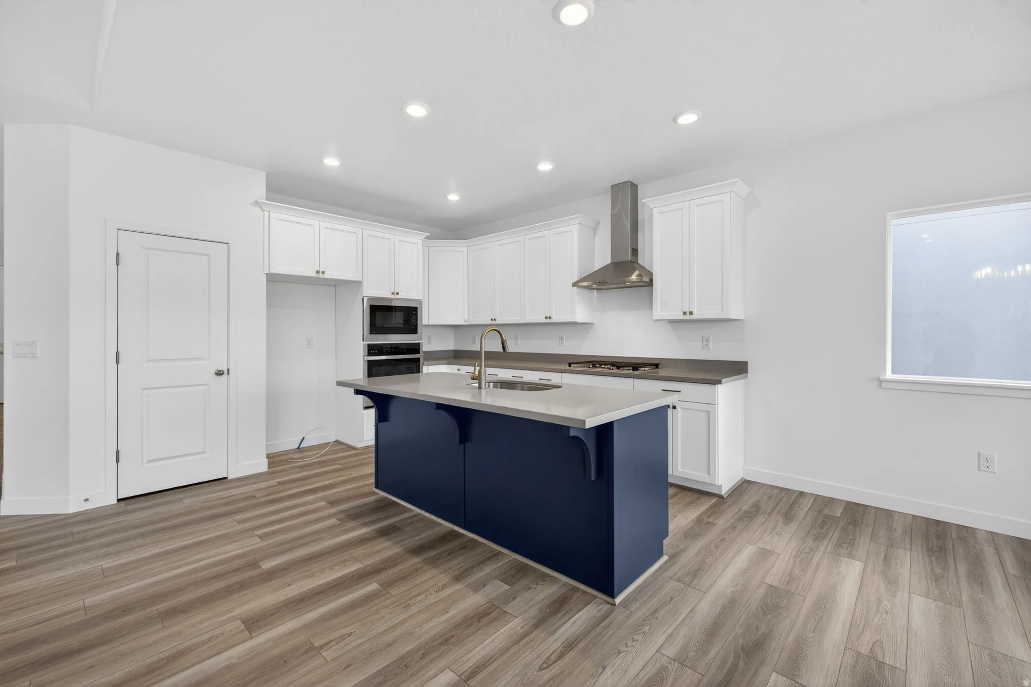 Kitchen featuring white upper and lower cabinetry, a blue kitchen island with a light-toned countertop, a stainless steel range hood, and wood-finish flooring