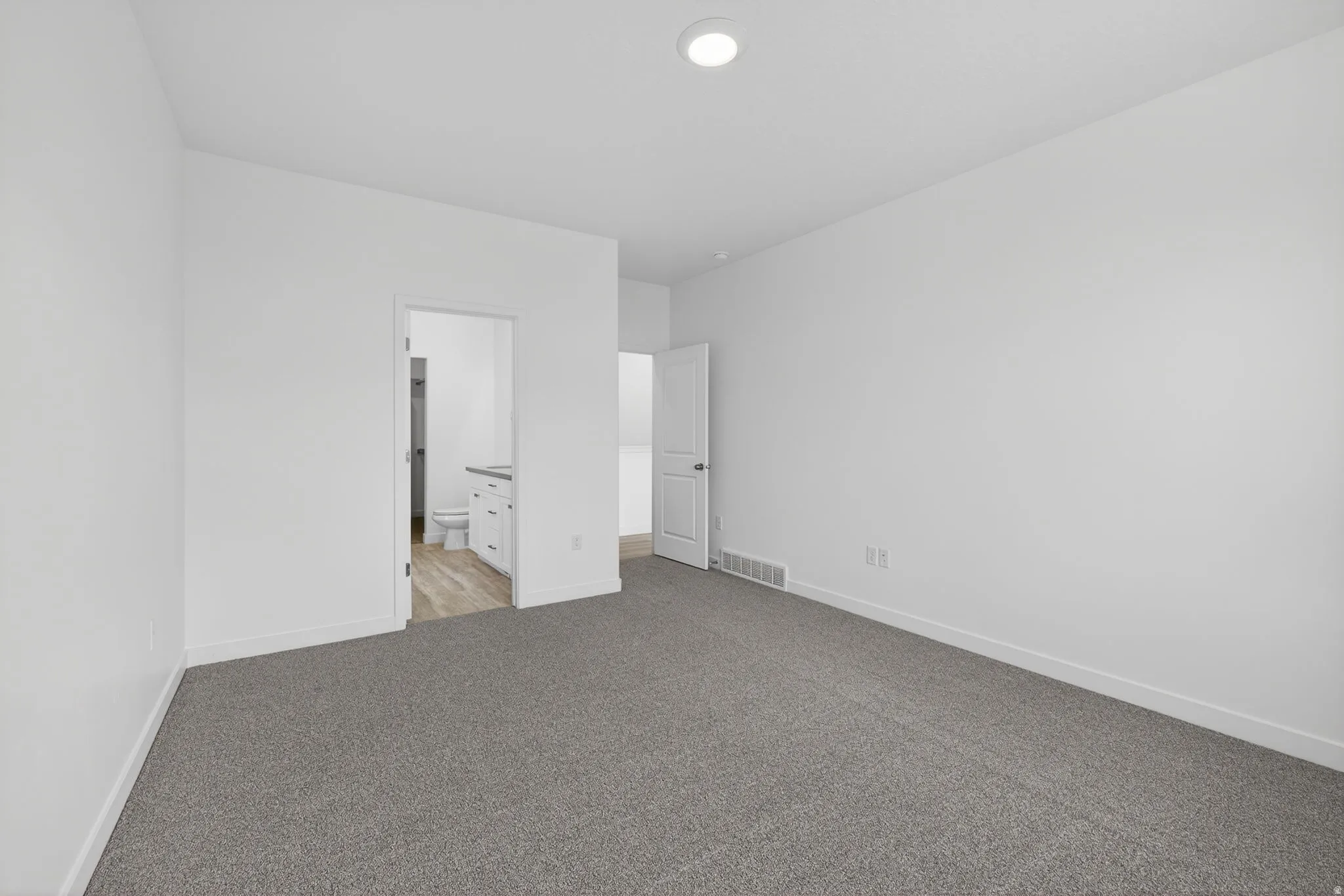 Plush grey carpeting, white interior walls, recessed ceiling light, white door, and an open doorway to a bathroom with wood-finish flooring and a vanity