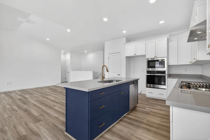 Kitchen featuring a blue island with integrated sink, stainless steel appliances including a wall oven and microwave, white cabinetry, and wood-finish flooring