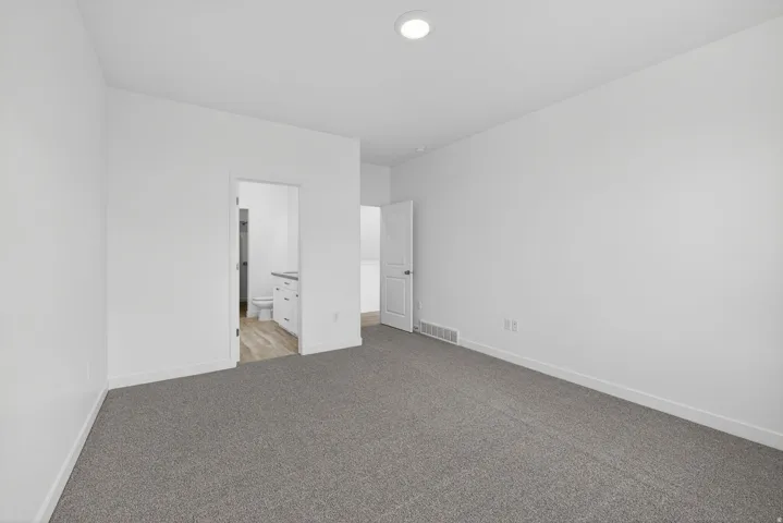 Plush grey carpeting, white interior walls, recessed ceiling light, white door, and an open doorway to a bathroom with wood-finish flooring and a vanity