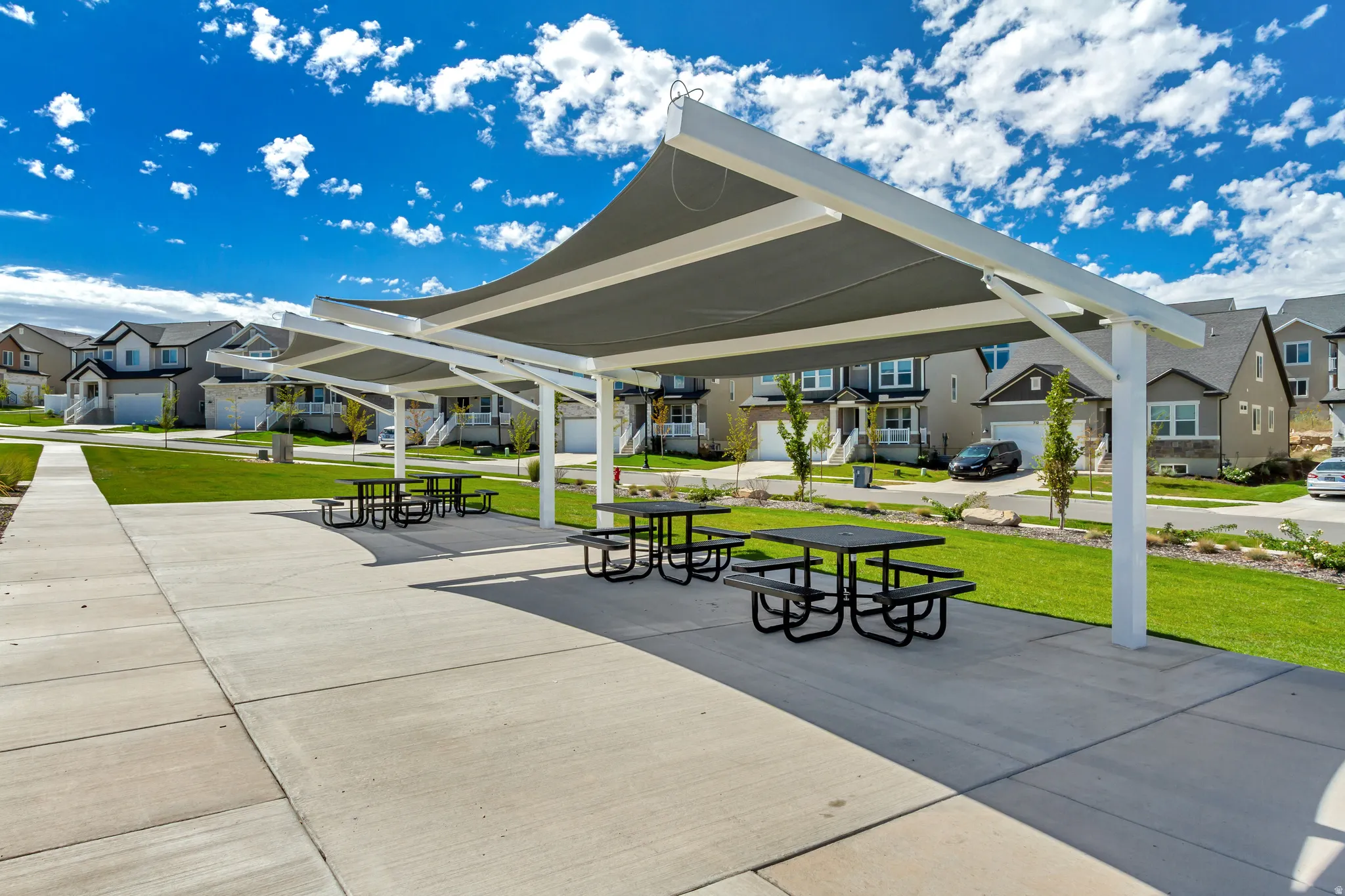 Community amenity area featuring multiple shaded picnic tables, expansive concrete paving, and a manicured lawn
